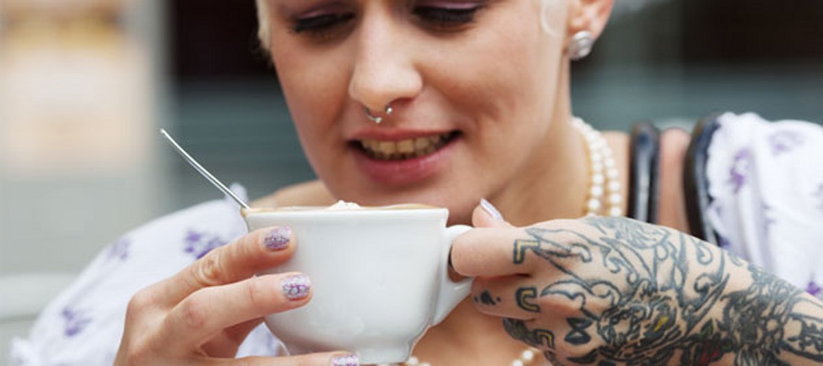 Eine junge Frau mit einer großen Tätowierung am Arm trinkt Kaffee.