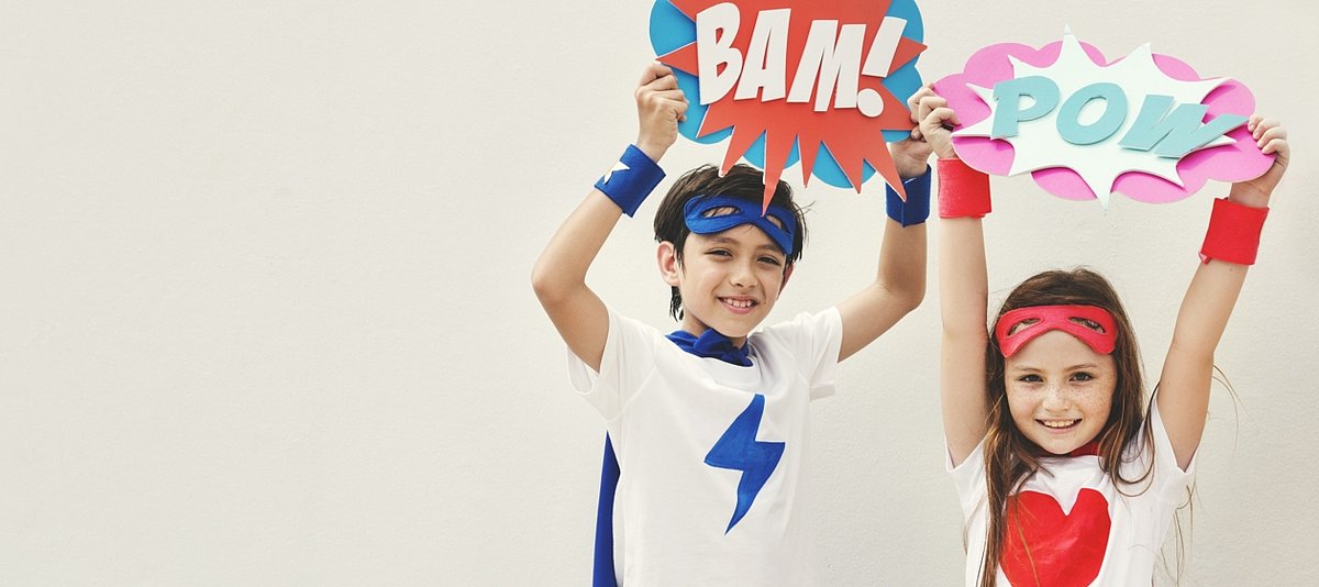 Ein Junge und ein Mädchen in Heldenverkleidung halten Schilder hoch mit der Aufschrift "Bam!" und "Pow"