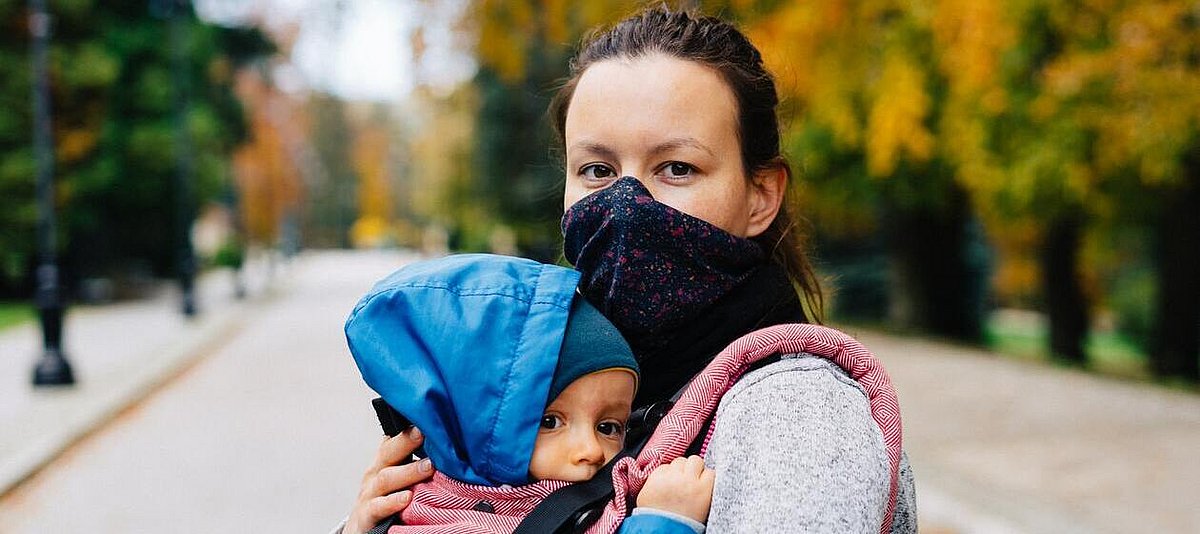 Frau mit Mundschutz trägt ein Baby mit Kapuze auf dem Arm