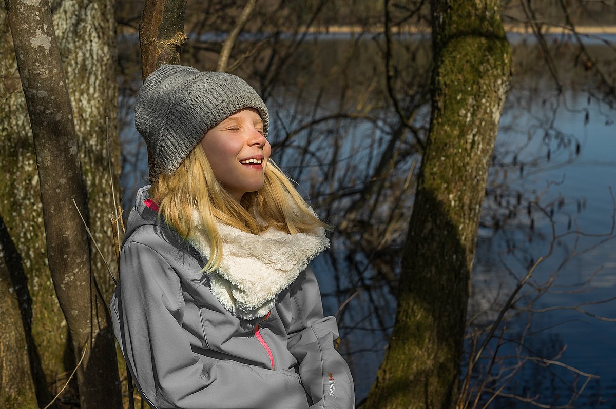 Eine Jugendliche lehnt an einem Baum und lächelt erleichternd, im Hintergrund ist ein See zu sehen
