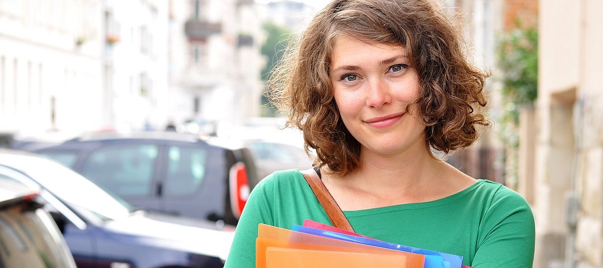 eine junge Studentin steht mit Heftern auf dem Fußweg und lächelt
