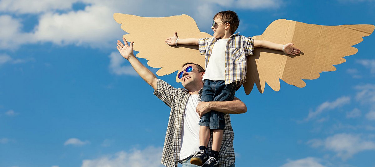 Ein Vater hat seinen Sohn, der Pappflügel trägt, auf dem Arm und beide tragen Sonnenbrillen und lachen.