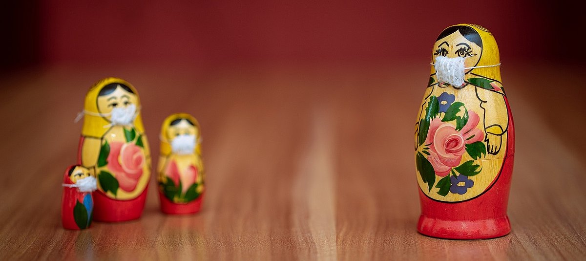 Drei kleinere Matrjoschka Figuren mit Mundschutz stehen links mit großem Abstand zu einer größeren Figur mit Mundschutz rechts im Bild 