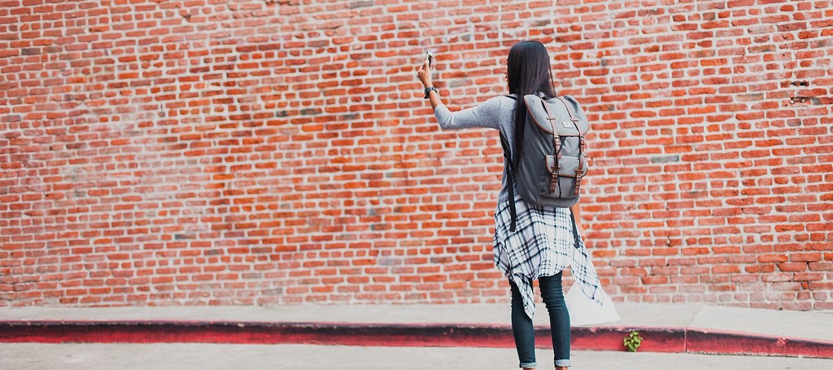 Eine junge frau mit Rucksack steht vor einer roten Wand und hält ihr Smartphone in der Hand 