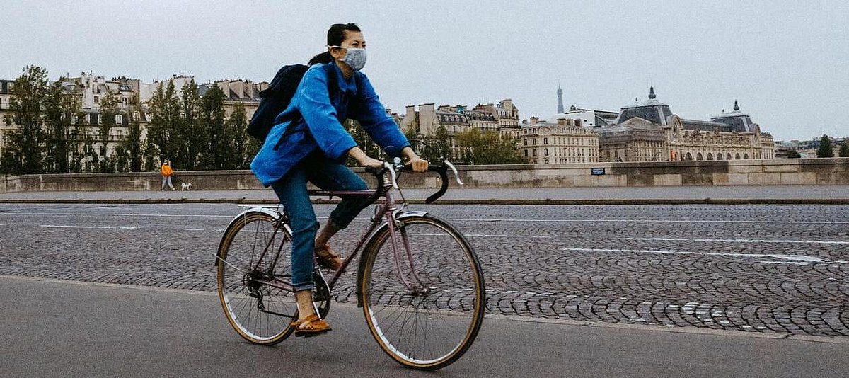 Eine junge Radfahrerin mit Mund-Nase-Schutz überquert auf dem Pont du Carrousel die Seine in Paris, im Hintergrund das Musée d'Orsay.