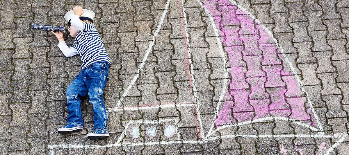 Ein Junge hat mit Kreide ein Boot auf die Straße gemalt und spielt Seemann.