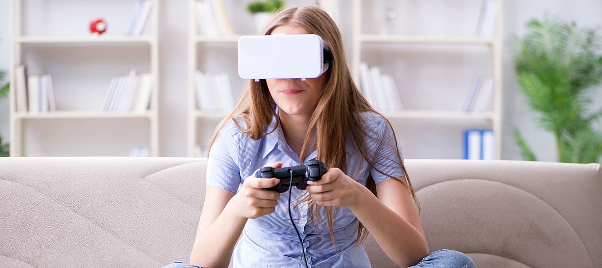 Eine Jugendlicher hat eine VR-Brille auf und sitzt spielend auf dem Wohnzimmersofa