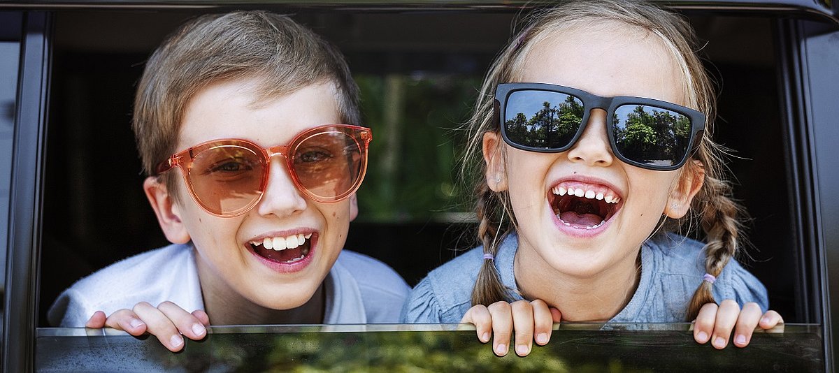 Zwei Kinder mit Sonnenbrille schauen aus dem heruntergelassenen Fenster eines Autos