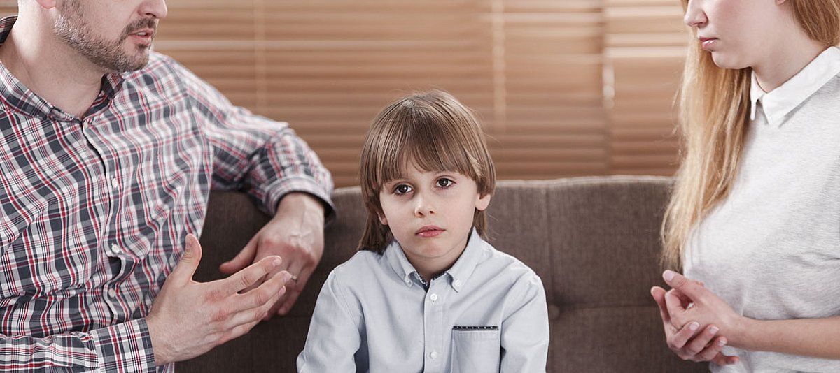 Eine Paar sitzt mit dem Kind in ihrer Mitte auf einem Sofa und führen ein sehr ernstes Gespräch, während der Junge sehr traurig schaut.