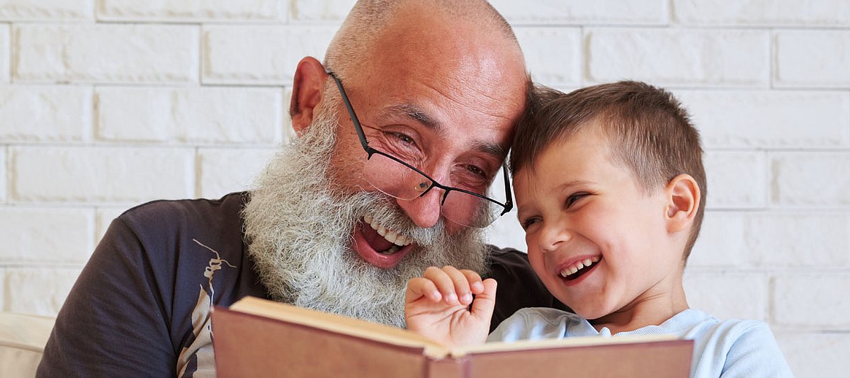 Ein Großvater liest mit seinem Enkel in einem Buch und beide lachen.