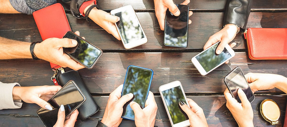 Erwachsene sitzen um einen Tisch und halten ein Smartphone in ihren Händen. 