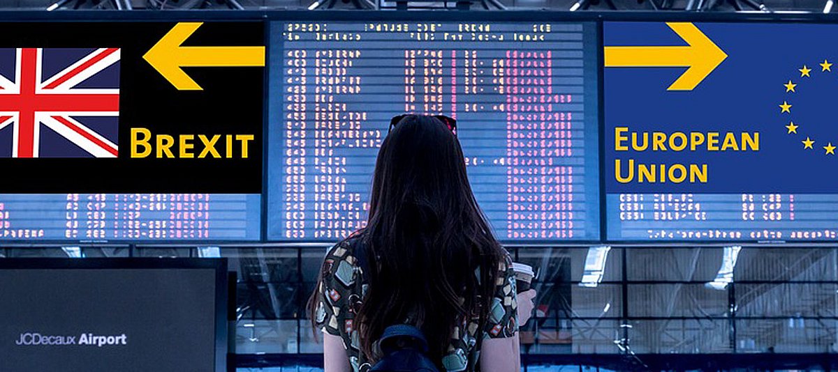 Eine junge Frau steht am Flughafen und schaut auf eine Informationstafel mit Fahnen von UK und EU mit Pfeilen nach Links und Rechts.