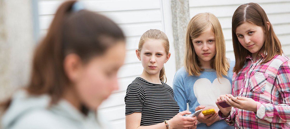 Drei Mädchen halten ihre Handys in der Hand und schauen verächtlich auf ein anderes Mädchen, das verschwommen im Vordergrund zu sehen ist