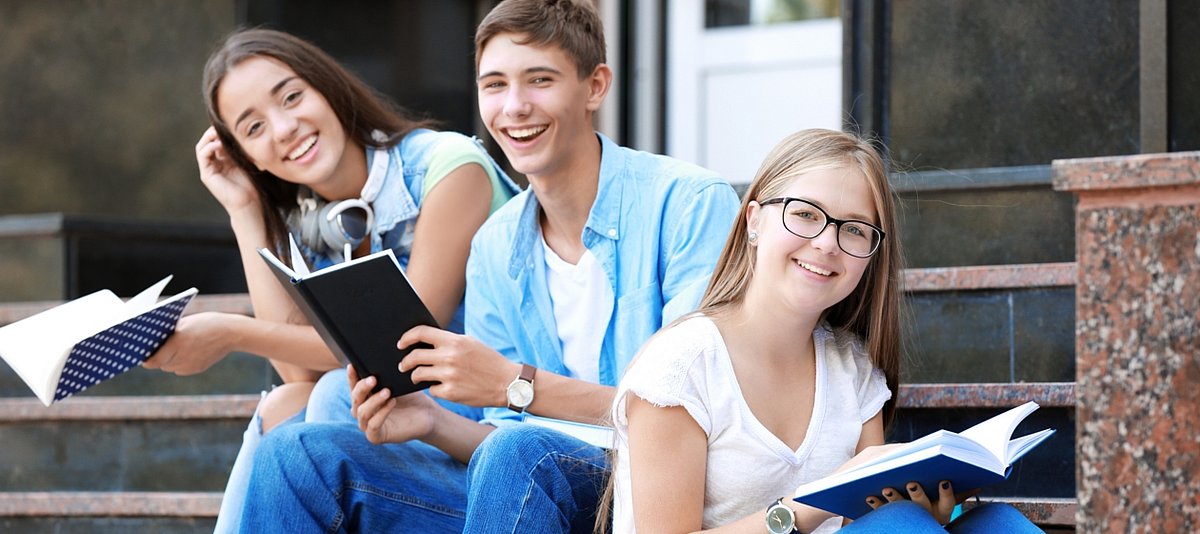 Drei Jugendliche sitzen vor der Schule auf einer Treppe, halten Hefte in der Hand und lachen