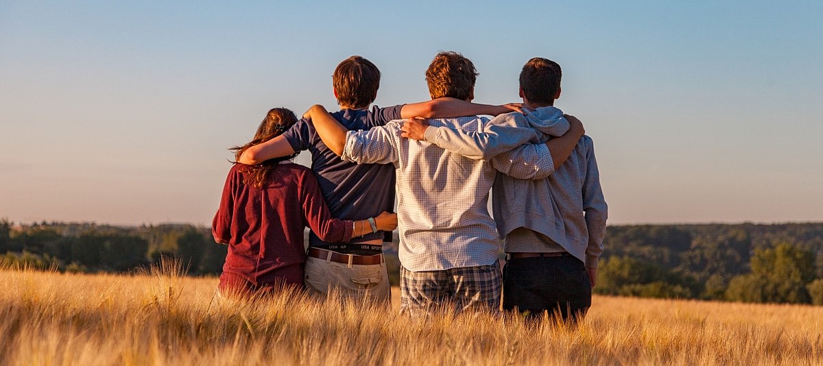 Vier junge Menschen stehen nebeneinander und mit Umarmung in der Abendsonne in einem Kornfeld