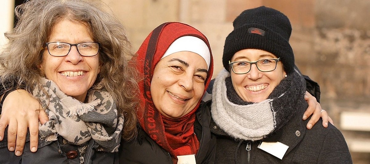 Drei Frauen umarmen sich lächelnd, die mittlere trägt ein Kopftuch
