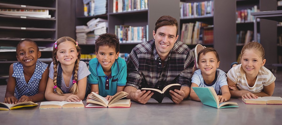 Mehrere Kinder und eine erwachsene Person liegen mit aufgeschlagenen Büchern vor sich in einer Bücherei