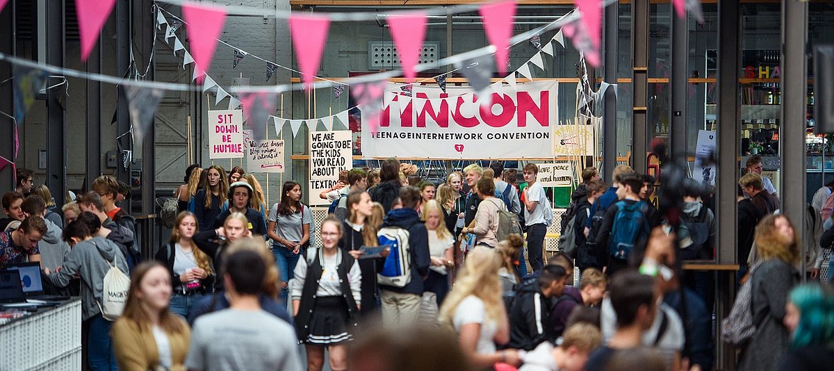 Viele junge Menschen in einem Veranstaltungsraum, pinkfarbenen Wimpelketten sind zu sehen und im Hintergrund das Logo der TINCON
