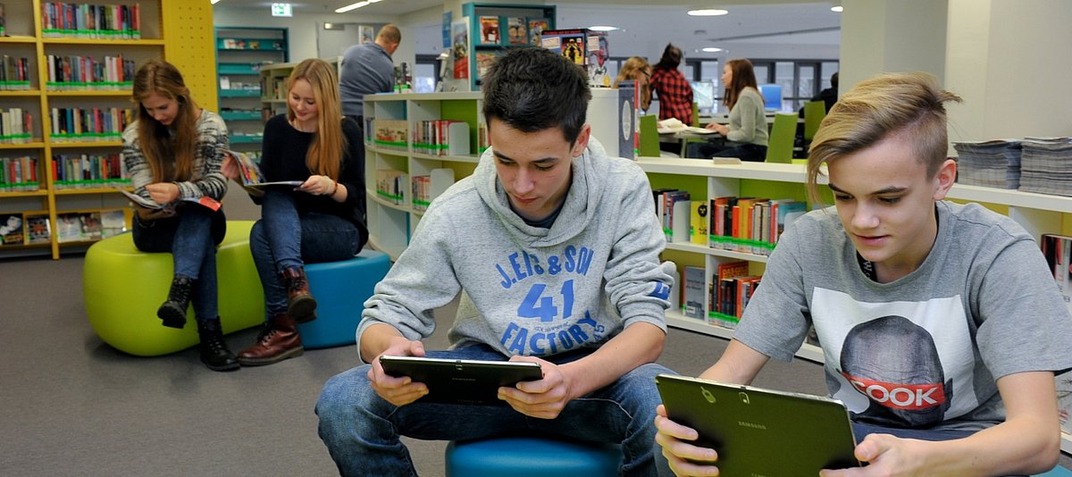 Schüler und Schülerinnen sitzen in einer Bibliothek und lesen auf Tablets