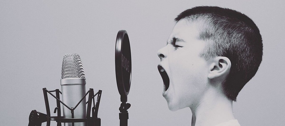 Ein Junge mit sehr kurzem Haar spricht mit aufgerissenem Mund in ein Studiomikrofon.