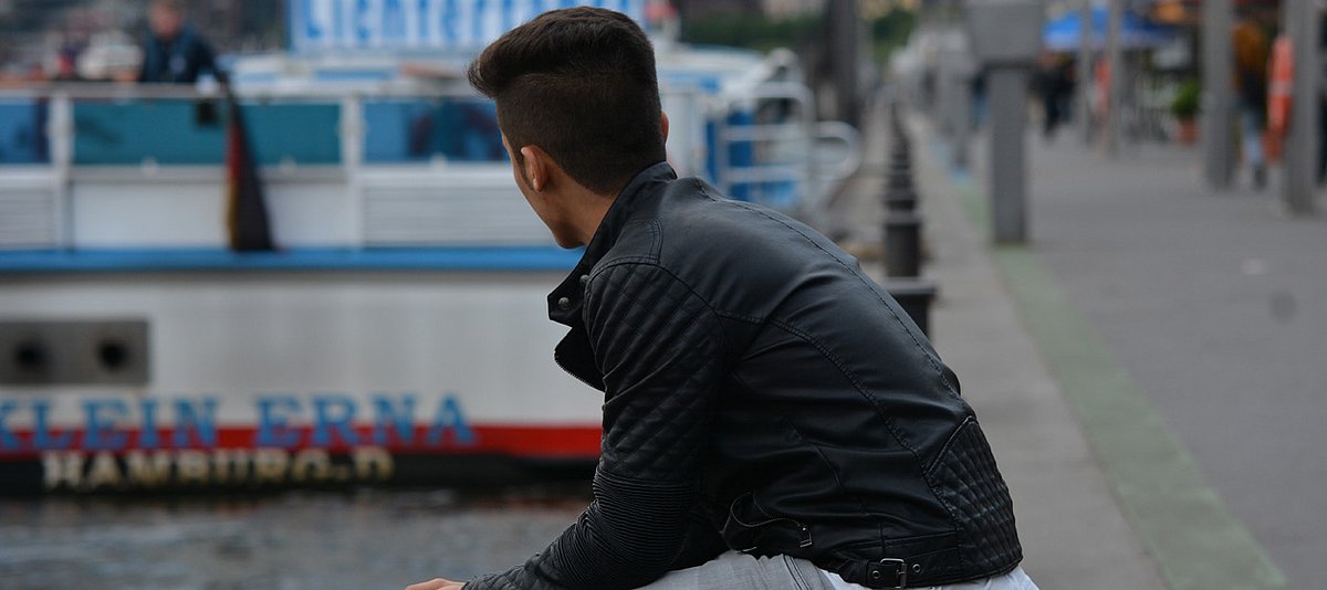 Ein Jugendlicher sitzt auf einer Kaimauer und schaut über das Hafenbecken. Im Hintergrund sieht man das Ausflugsschiff "Klein-Erna" und ein Stück der Hafenpromenade 