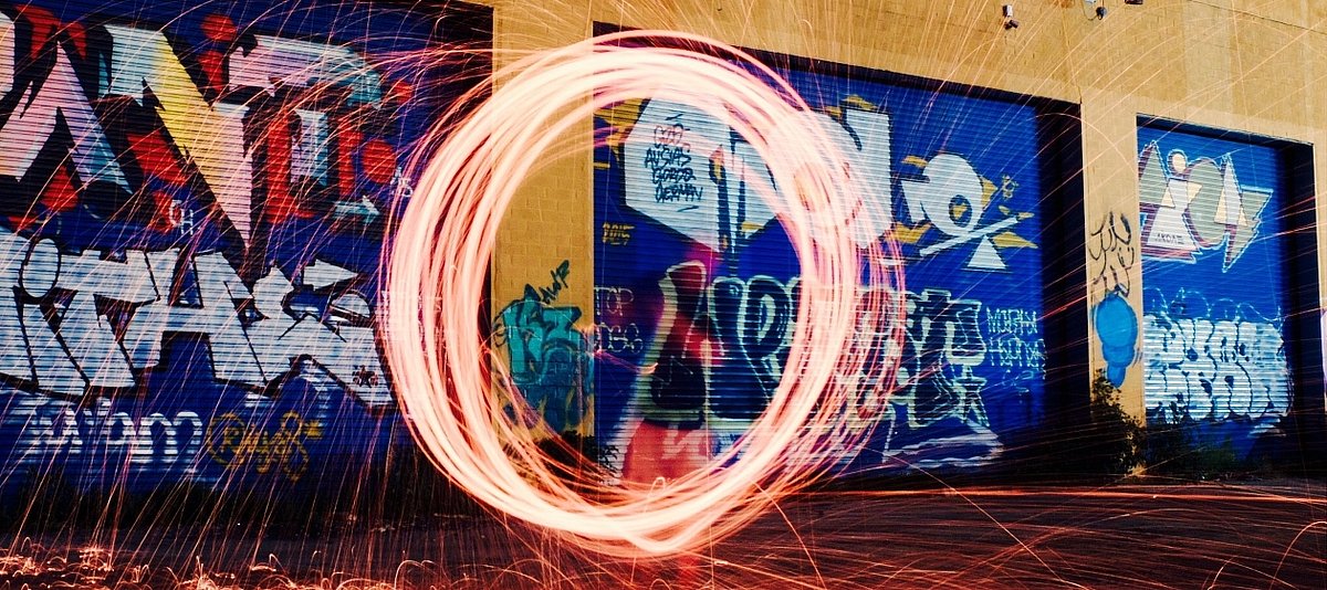 Eine Person steht vor einer Wand mit Graffittis und durch eine Langzeitbelichtung ist ein mit einer Lichtquelle in die Luft gemalter Kreis zu sehen