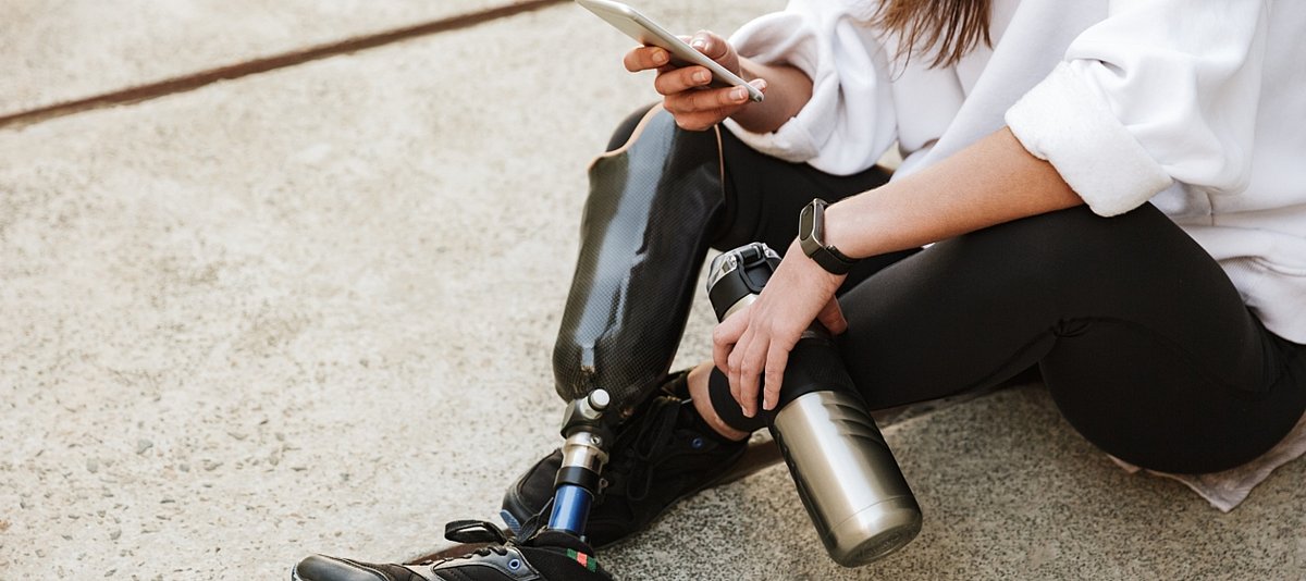 Frau mit Beinprothese sitzt auf dem Boden und tippt in ihr Handy