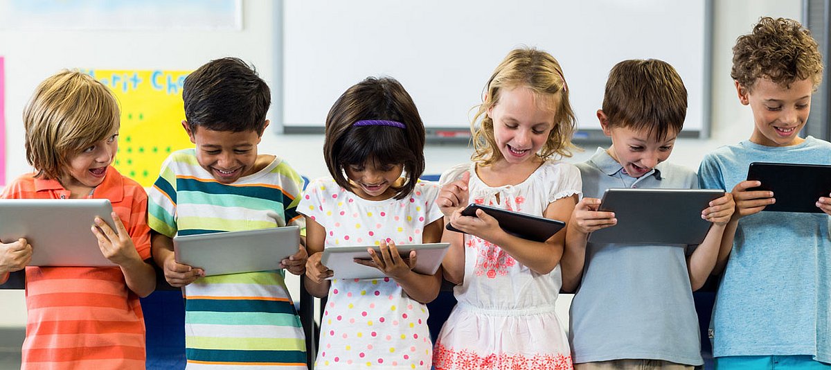 Sechs Kinder im Grundschulalter, unterschiedlicher Herkunft, stehen mit Tablets in den Händen in der Schule und lachen. 