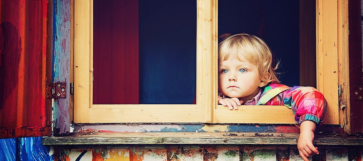 Ein Kind mit einem bunten Oberteil schaut aus einem Fenster mit Fensterläden