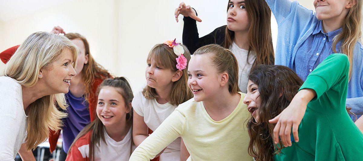 Ein Gruppe von Schülerinnen im Teenageralter üben mit einer Lehrerin Mimik und Gestik. 