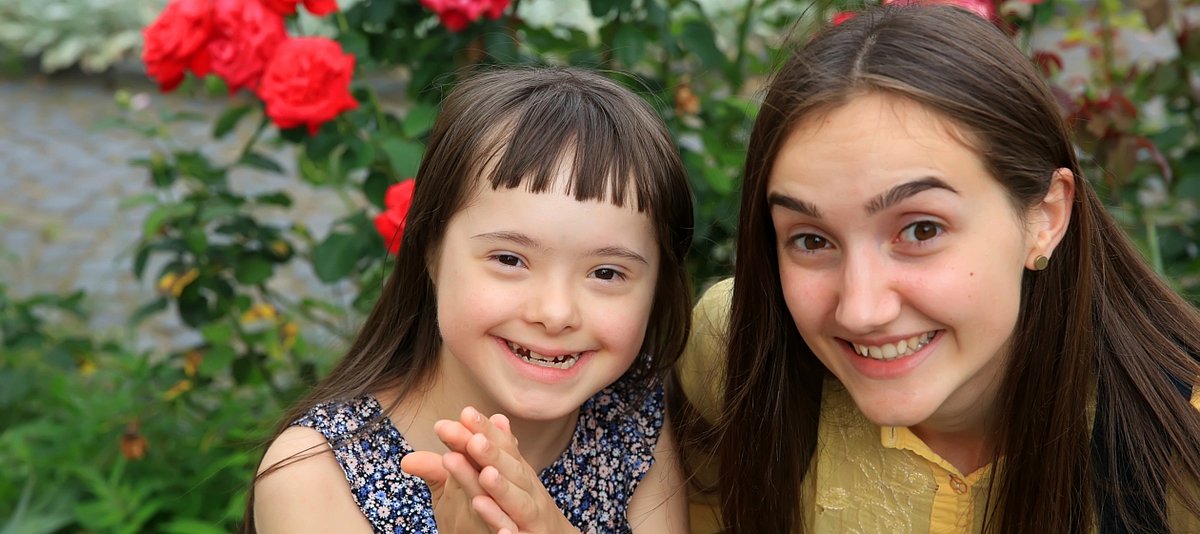 Mädchen mit Downsyndrom lächelt zusammen mit älterem Mädchen in die Kamera