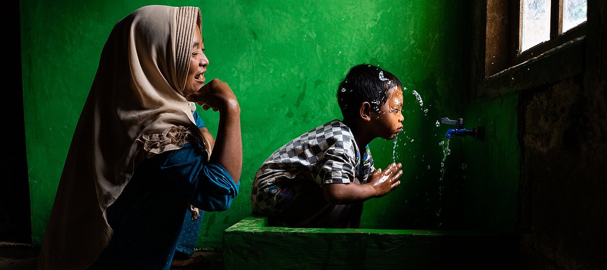 Indonesisches Kind spielt am Waschbecken mit Wasser und wird von Mutter beobachtet.