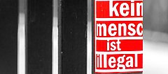 Auf dem Bild ist ein Tor mit einem roten Sticker und weißer Aufschrift "Kein Mensch ist illegal" zu sehen