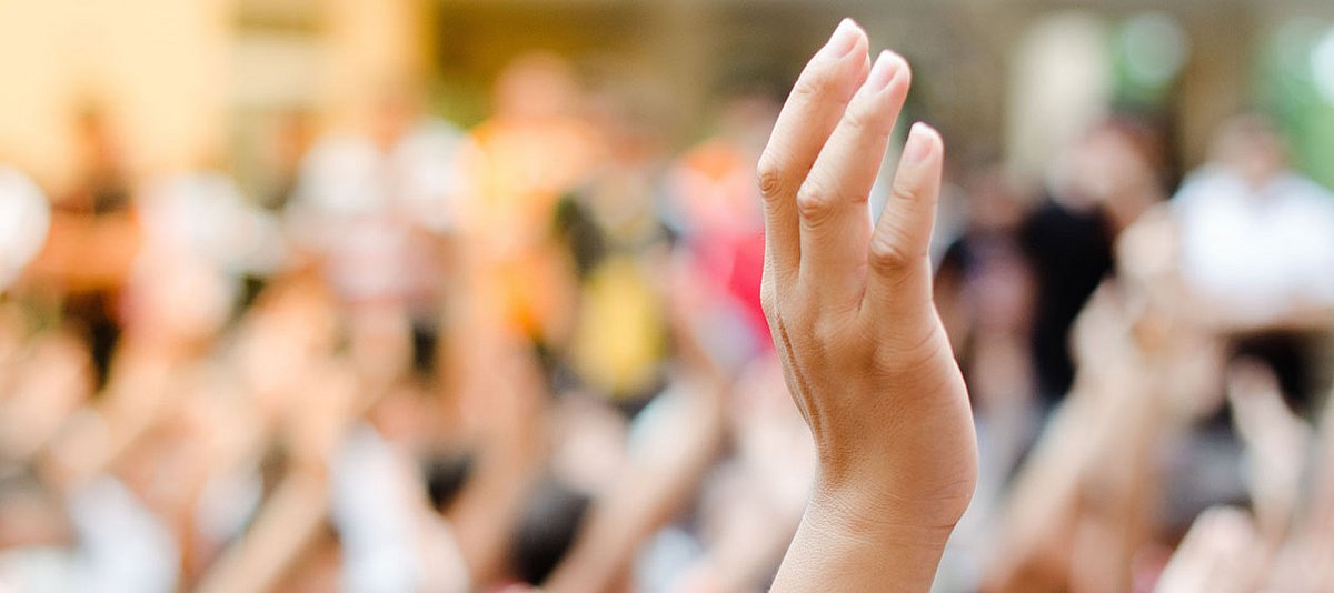 Eine Hand ist erhoben vor dem Hintergrund einer Gruppe mit erhobenen Händen zu sehen.