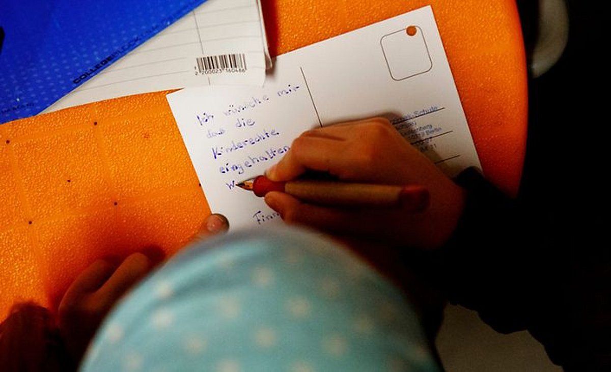 Kinderhand mit Füller schreibt auf Postkarte: "Ich wünsche mir, das die Kinderrechte eingehalten w... Finn"