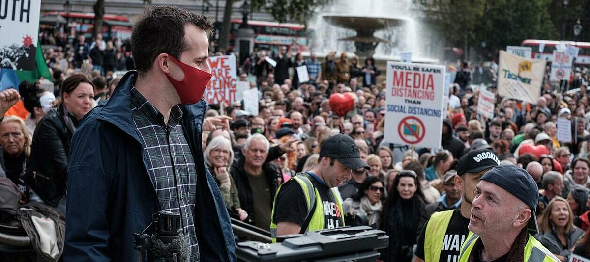 Ein Journalist mit rotem Mund-Nase-Schutz steht auf einem Platz mit Springbrunnen vor einer großen Menge von Anti-Corona-Demonstranten, fast alle ohne Maske.