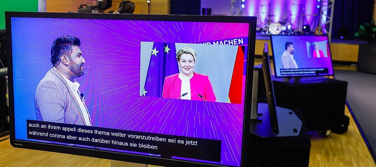 Ein violetter Bildschirm mit bärtigem Moderator links und Bundesministerin Giffey, zugeschaltet, vor den Flaggen der BRD und der EU.