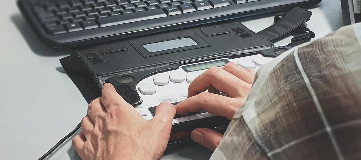 Ein Mann tippt auf einer Brailletastatur. Dahinter liegt eine Standardtastatur.
