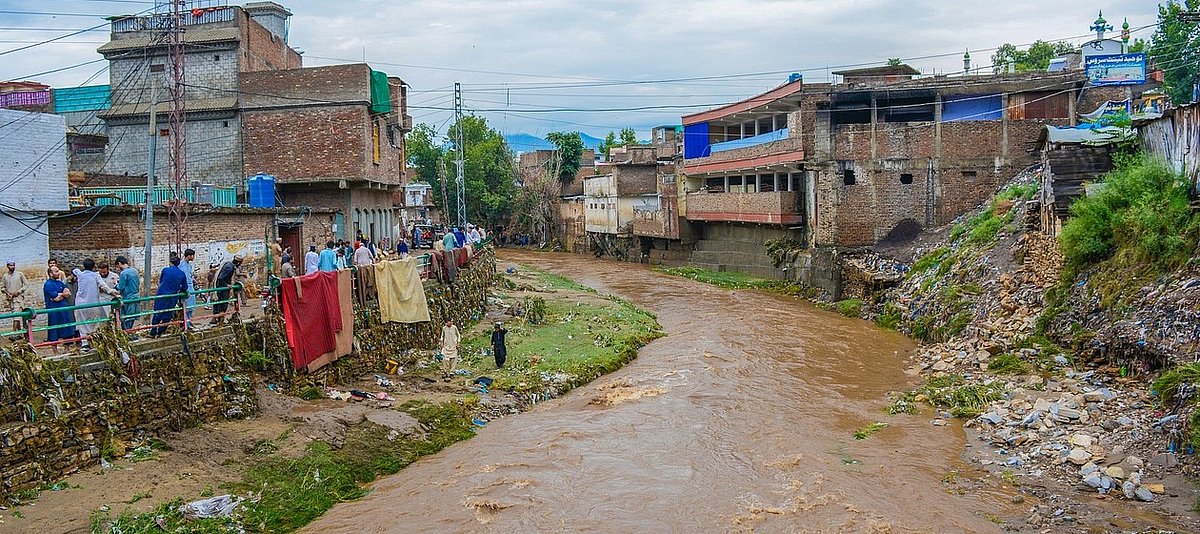 Auf dem Bild ist ein schlammiger Fluss und einige Häuser im Swap-Bezirk in Pakistan zu sehen, die Häuser sind teils zerstört sind von den Überflutungen 