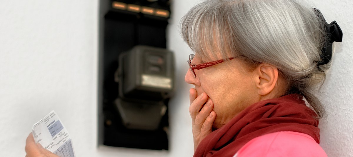 Eine Frau steht vor dem Stromzähler und blickt mit Hand vor dem Mund auf eine Rechnung. 