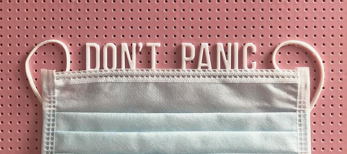 In einer rosa Steckwand steckt der Buchstabenzug DON'T PANIC, darunter eine hellblaue OP-Maske.