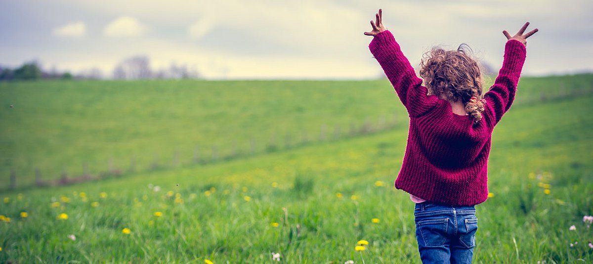Ein Mädchen im Kindergartenalter steht auf einer großen Wiese und streckt die Hände glücklich in die Luft.