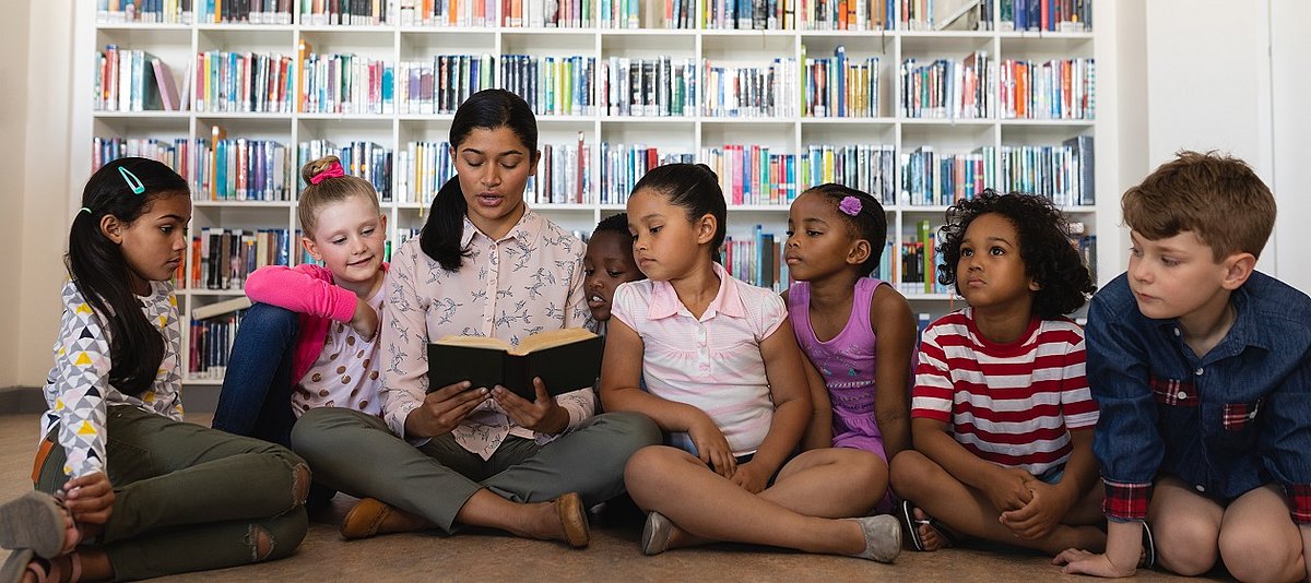 Eine erwachsene Person liest einer Gruppe Kindern vor, im Hintergrund ist ein Bücherregal