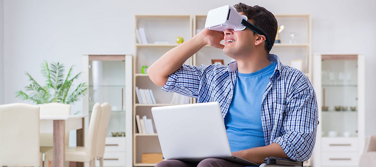 Ein junger Mann sitzt im Rollstuhl im Wohnzimmer und schaut mit dem Laptop auf den Beinen in eine Virtual Reality Brille.
