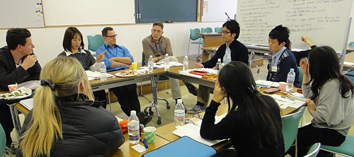 Deutsche und japanische Teilnehmende bei einem Fachkräfteaustausch sitzen an einem Tisch in einem Seminarraum. Im Hintergrund sieht man eine viel beschriebene Tafel.