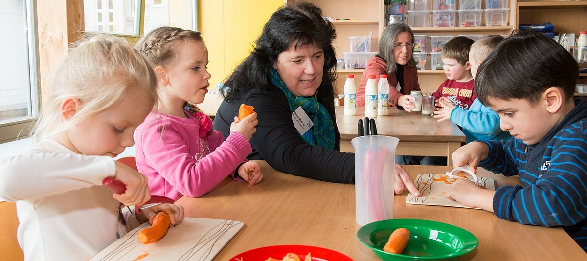 Eine Erzieherin sitzt mit Kindern an einem Tisch, die Gemüse schälen