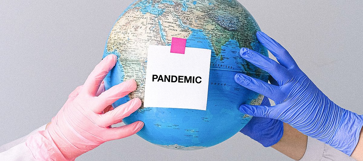 Zettel mit dem Wort Pandemic aufgeklebt auf einen Globus, der von vier Händen in Gummihandschuhen gehalten wird