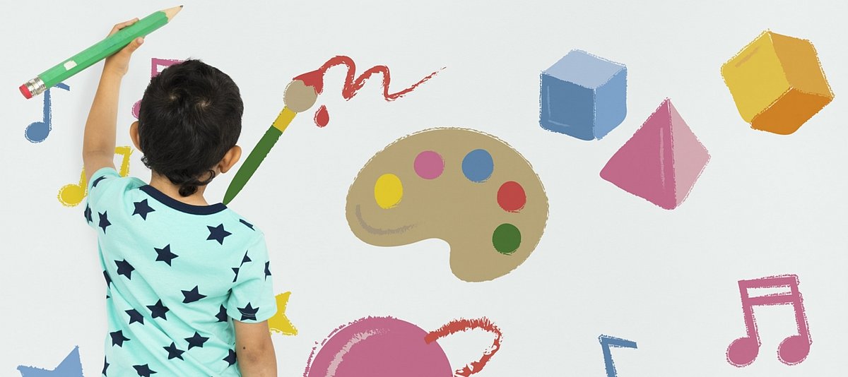 Ein Junge steht vor einer Wand mit Abbildungen von u.a. einer Farbpalette, Bauklötzen und Musiknoten, erselbst hält einen großen Bleistift in der Hand und schreibt etwas