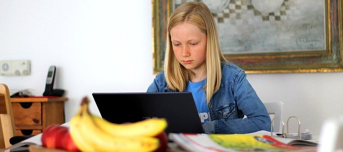 Ein blondes Mädchen in Jeansjacke sitzt am Küchentisch, auf dem Bananen und anderes Obst liegen, am Laptop und ist mit Schulaufgaben beschäftigt