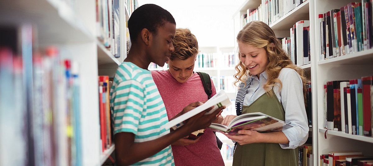 Drei Jugendliche stehen zwischen Bücherregalen und lesen in Büchern